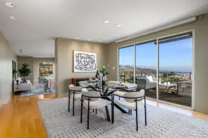 Real estate Photograph of a bright and spacious dining area featuring a sleek black dining table, elegant white chairs, and large sliding glass opening to a balcony with panoramic city views of the Hollywood Hills .